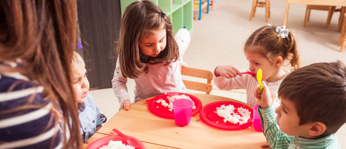 Glutenfrei in Kita und Schule Kitakinder sitzen während des Essens gemeinsam am Tisch. Pädagogische Fachkraft unterstützt die Kinder dabei.